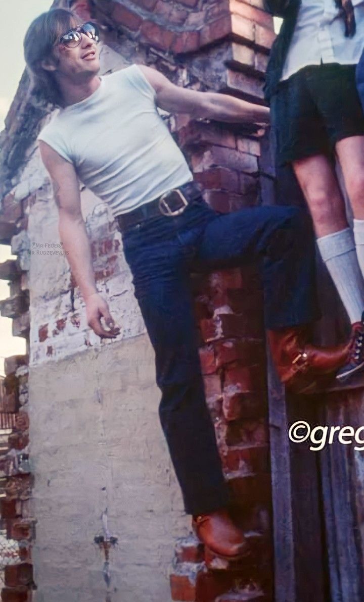 two men standing on the side of a brick building with their legs hanging out and one man wearing glasses