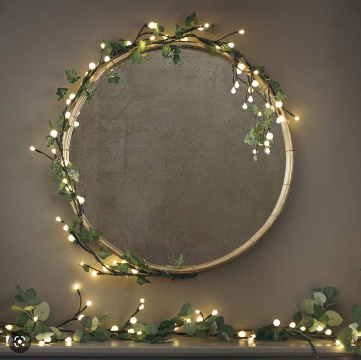 a round mirror with lights around it on a mantle next to a wreath and ivy garland