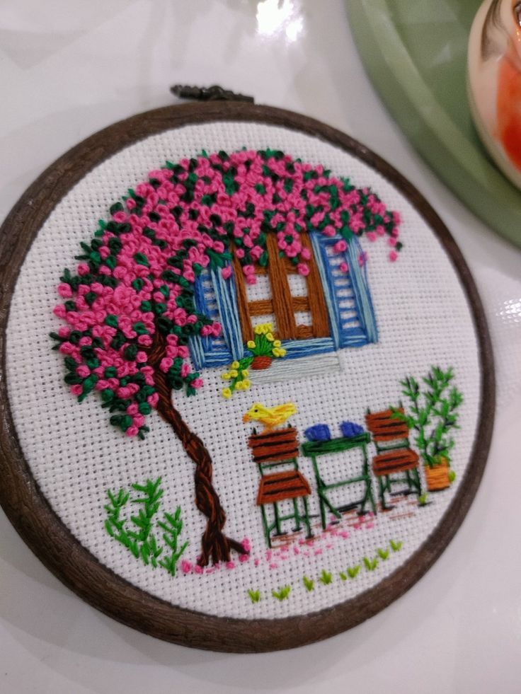 a cross stitched picture of a table and chairs under a tree with pink flowers