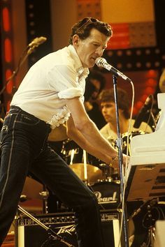 a man standing in front of a white piano on stage with other musicians behind him