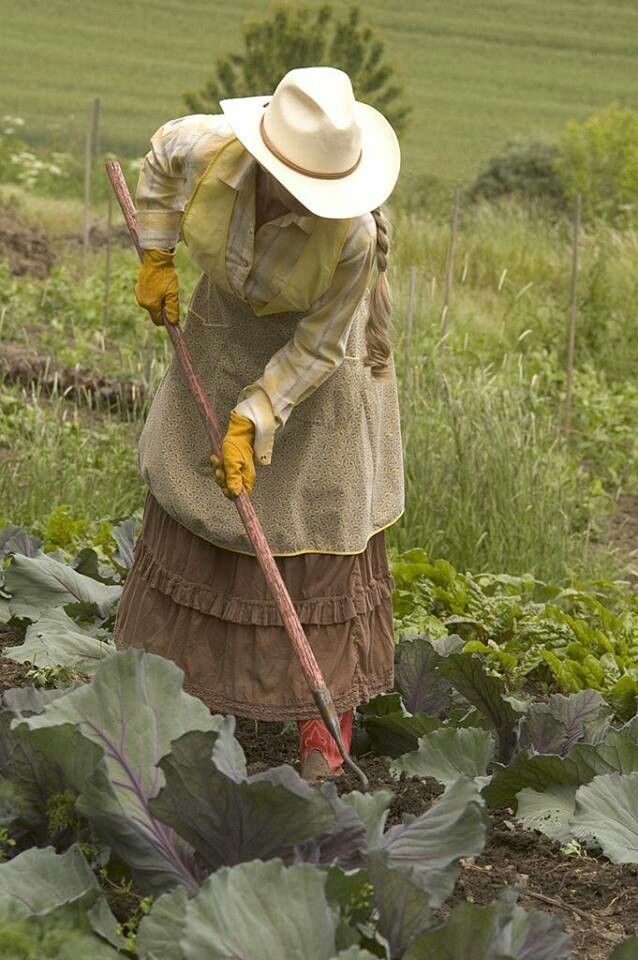 a woman in a hat and dress is working in the garden with a stick on her shoulder