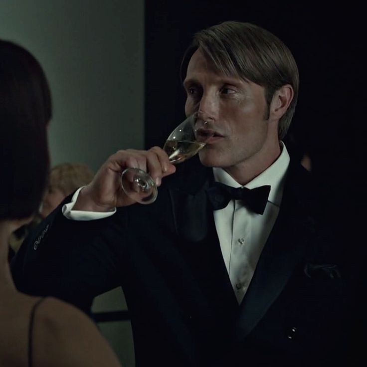 a man in a tuxedo drinking from a wine glass while standing next to a woman