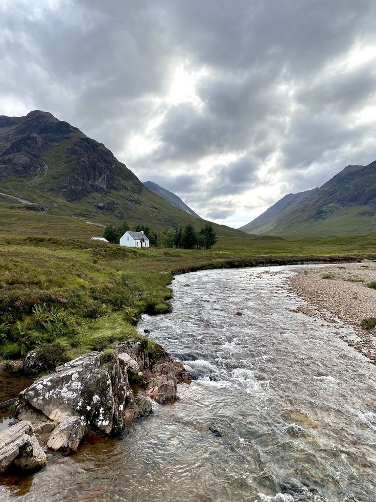 a river running through a lush green valley under a cloudy sky with mountains in the background