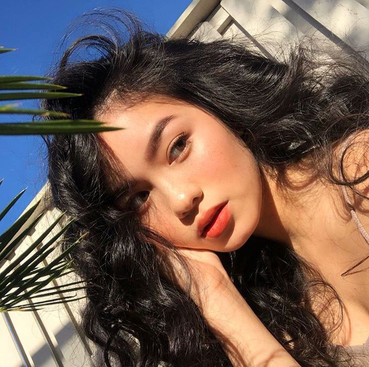 a woman with long black hair laying on the ground next to a palm tree and looking off into the distance