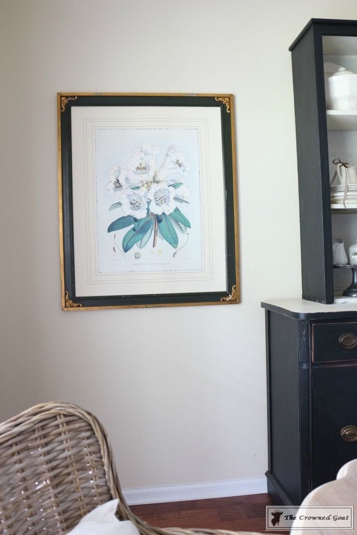 a painting hangs on the wall next to a wicker chair and china cabinet in a white walled room