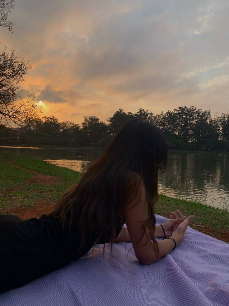 a woman laying on top of a blanket next to a body of water at sunset