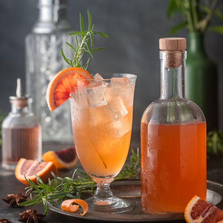 a bottle of alcohol next to a glass with an orange slice and rosemary garnish