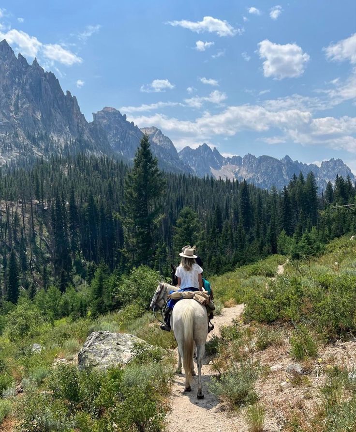 a person riding on the back of a white horse down a dirt road in front of mountains