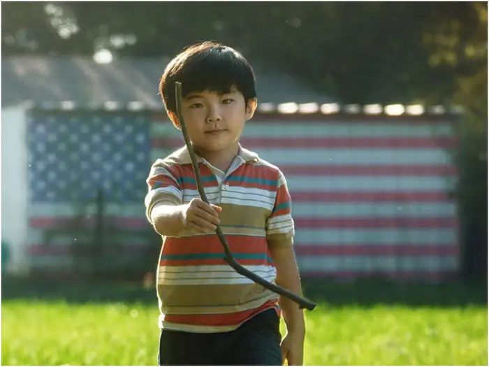 a young boy holding a hose in his hand while standing on the grass with an american flag behind him