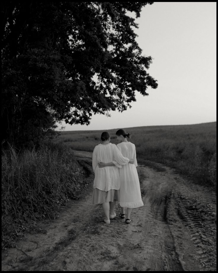 two women in white dresses walking down a dirt road next to a large leafy tree