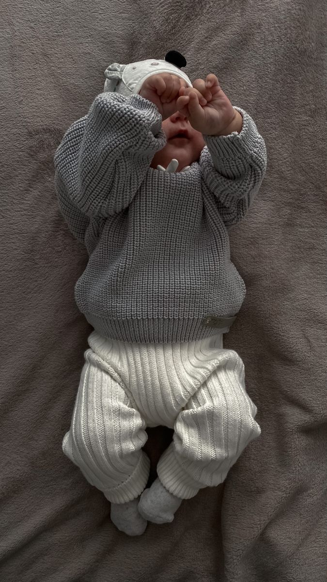 a baby laying on top of a bed wearing a gray sweater and white leggings