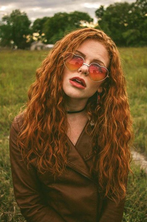 a woman with long red hair and glasses sitting on the ground in a grassy field