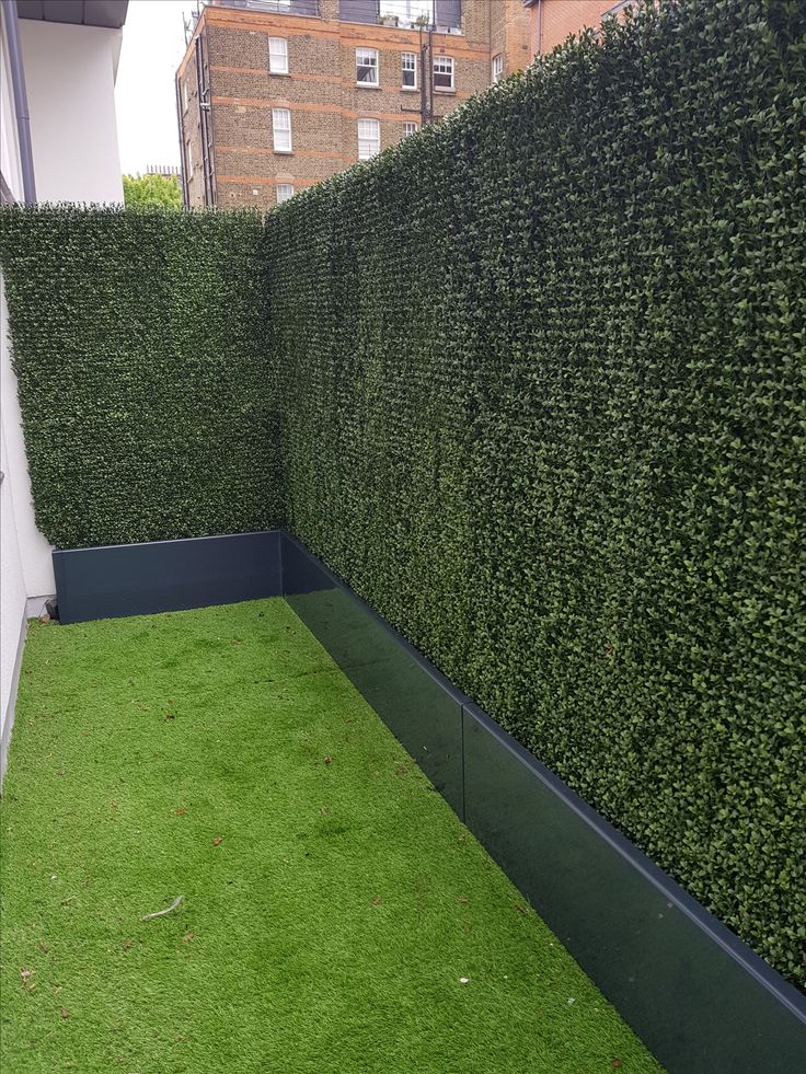 an artificial grass area in front of a building with a large green hedge on the side