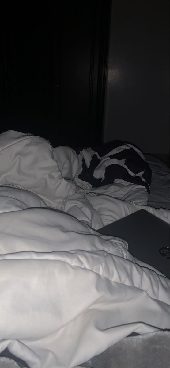 an unmade bed with white sheets and blankets on it in the dark, next to a laptop computer