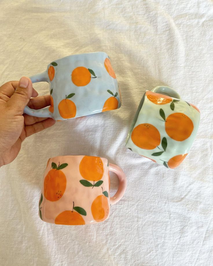 three oranges painted on ceramic mugs sitting on a white sheet, one being held up by a hand