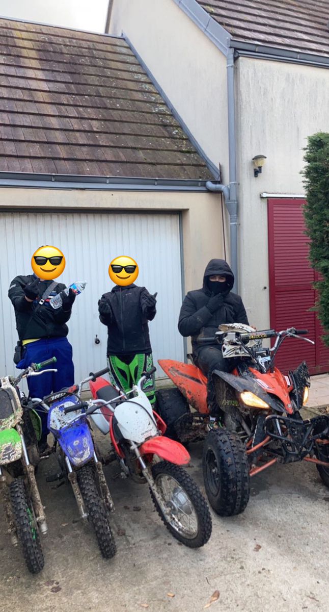 three people wearing helmets and standing next to four dirt bikes in front of a garage