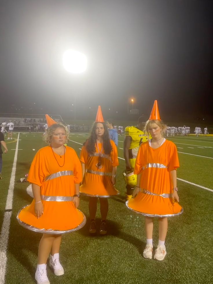 three girls in orange dresses standing on a football field at night, wearing party hats