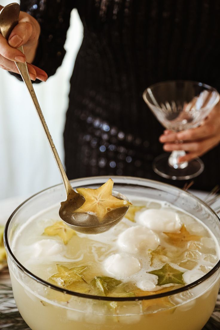 a woman is spooning ice into a bowl filled with fruit and vegetables, while another person holds a wine glass in the background