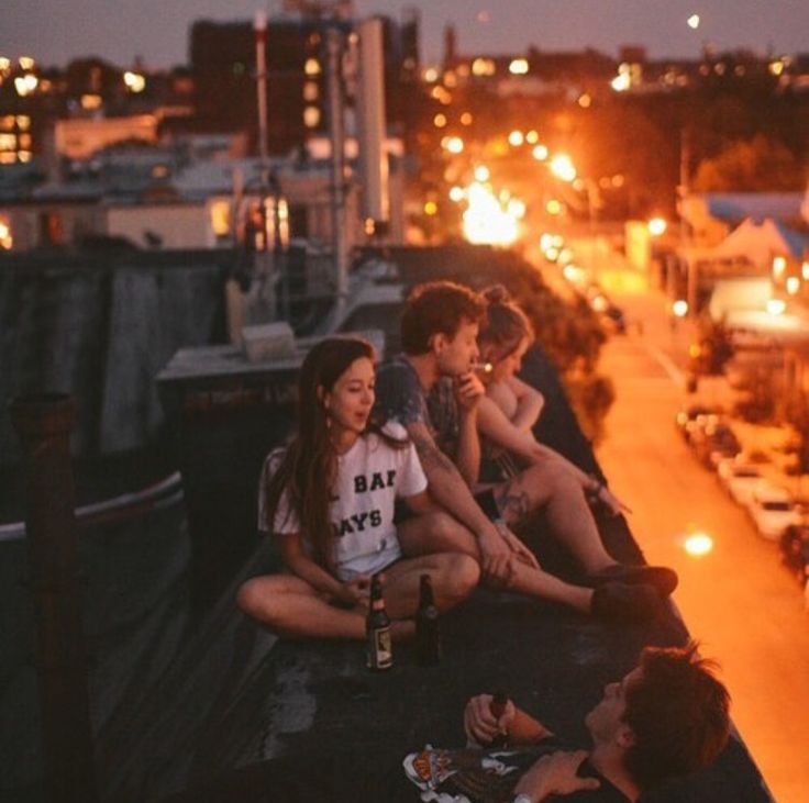 four people sitting on the edge of a bridge at night eating and drinking beer,