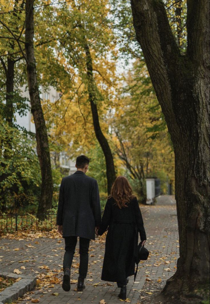 a man and woman holding hands walking down a sidewalk in the fall with leaves on the ground