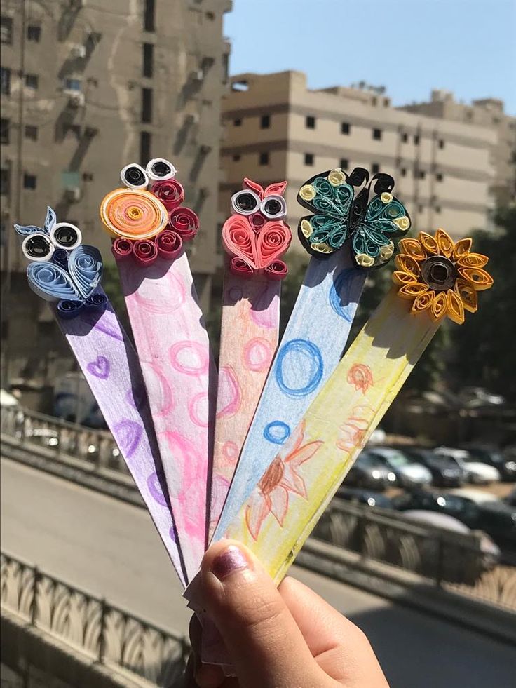 a hand holding five colorful paper flowers in front of a cityscape with buildings