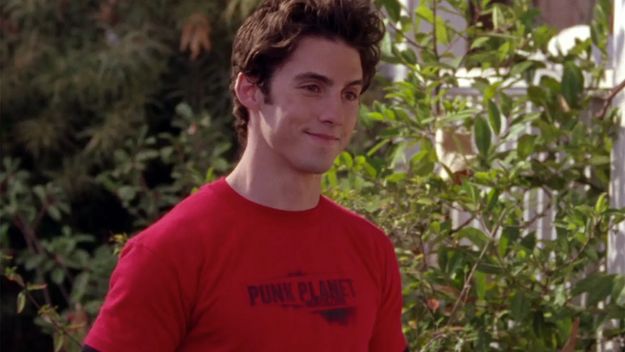 a young man in a red shirt standing next to some trees and bushes with his hand on his hip