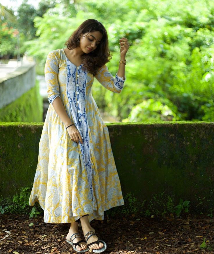 a woman in a yellow and blue dress is standing by a green wall with her hand on her hip