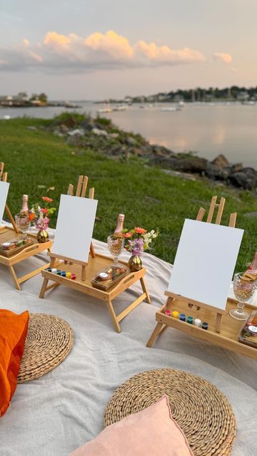 an outdoor setting with easels and paintings on the grass by the water at sunset