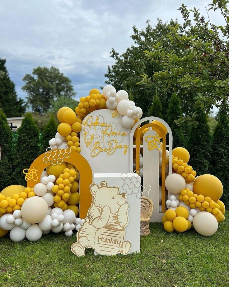 a yellow and white birthday party with balloons on the grass, decorations and trees in the background