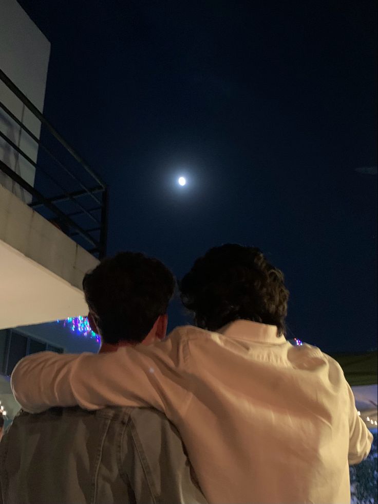 two people are sitting on a balcony watching the moon go down in the sky above them