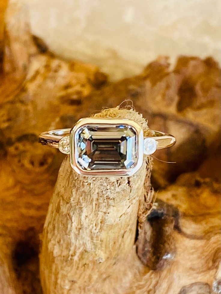 This classic east west bezel setting contains a beautiful 2.00ct. emerald cut gray moissanite with 1.7mm round moissanite accents. The center stone is VVS clarity, measures 8x6mm and comes with a grading certificate. The ring is available in sterling silver, and 14k white, yellow, or rose gold.  This ring makes a great promise or engagement ring.  Please send me a message if you need a size not listed.  *  This ring can be customized with any color center stone. All items are handmade by me in m Silver Diamond Ring With Radiant Rose Cut, Sterling Silver Emerald Cut Diamond Ring With Bezel Setting, Horizontal Emerald Engagement Ring Bezel, Silver Emerald-cut Ring With Rose Cut Diamonds, East West Emerald Bezel Engagement Ring, East To West Emerald Engagement Ring, Emerald East West Engagement Ring, Sterling Silver Diamond Ring With Bezel Setting, Emerald Cut, Emerald Cut East West Engagement Ring