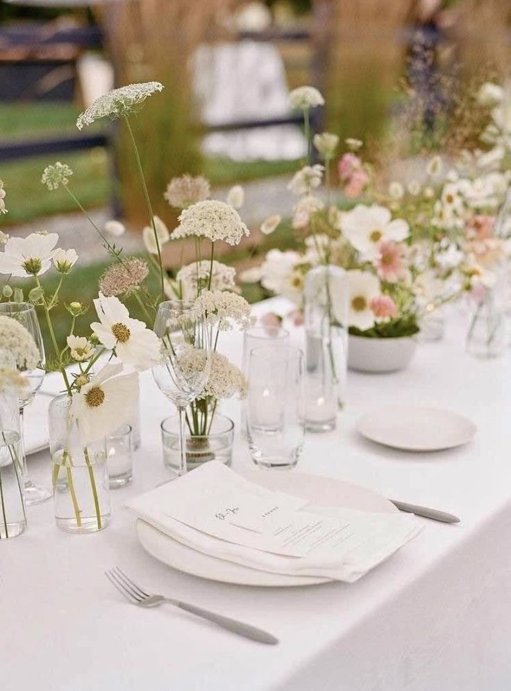 the table is set with white and pink flowers in glass vases, plates and silverware