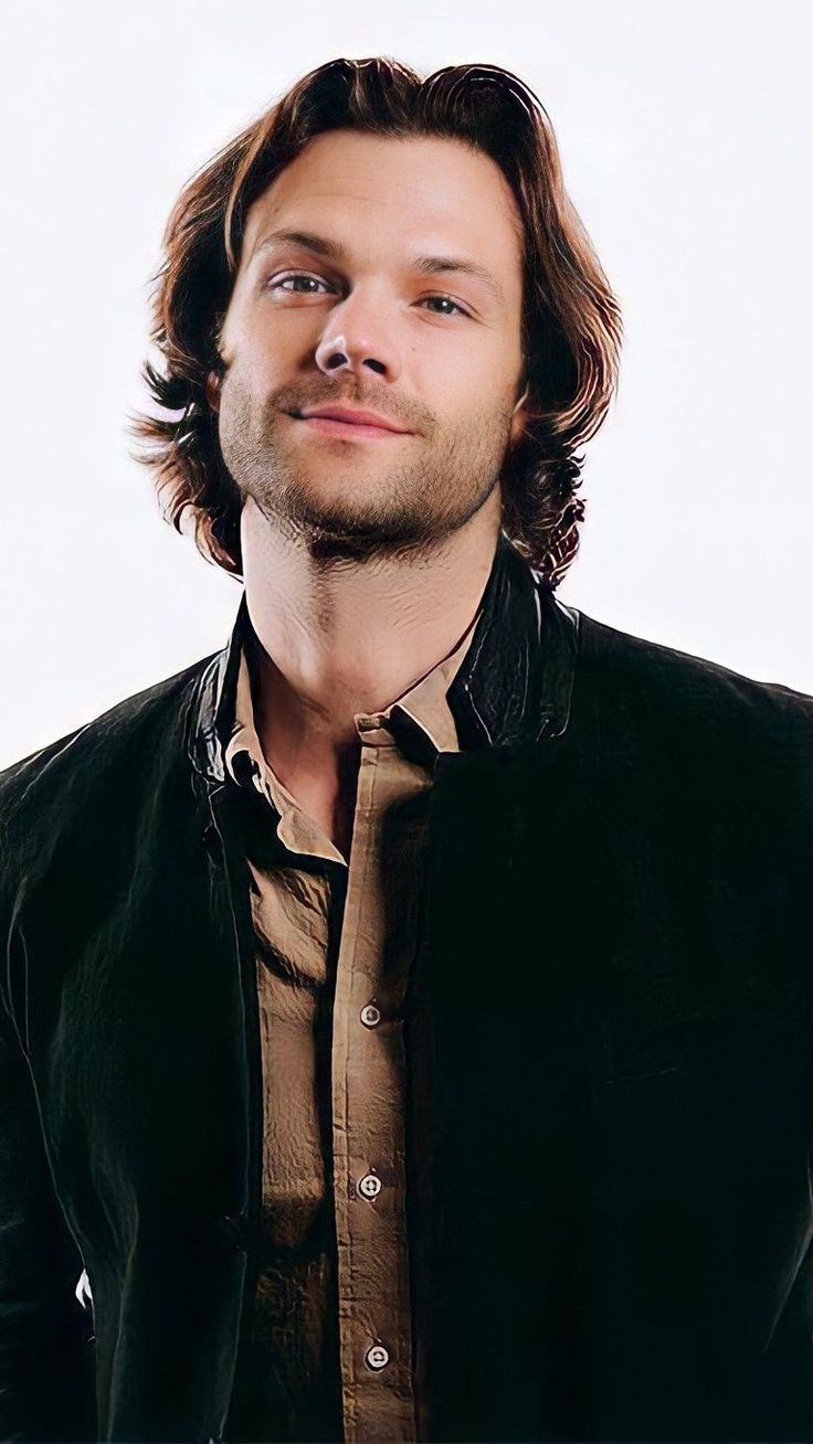 a man with long hair wearing a black jacket and looking to the side while standing in front of a white background