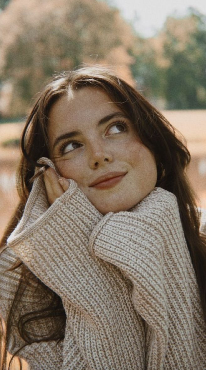 a woman with long brown hair wearing a sweater and holding her hand on her face