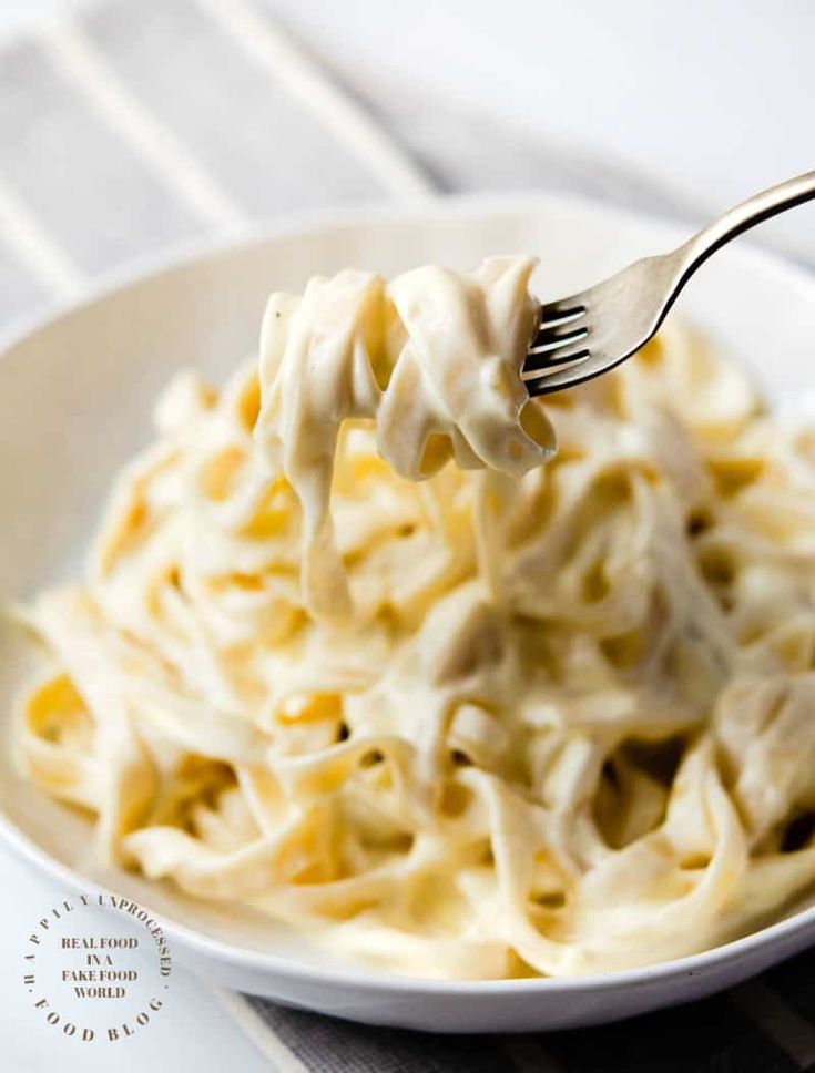 a fork full of pasta with cream sauce on it, in a white bowl that is sitting on a table