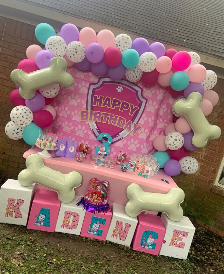 a birthday cake table with balloons and dog bone decorations on the top, in front of a brick wall