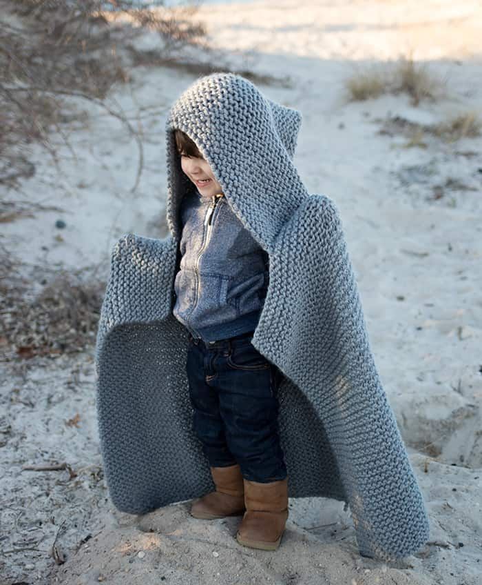 a little boy standing in the sand wearing a knitted hooded jacket and boots with his arms wrapped around an object