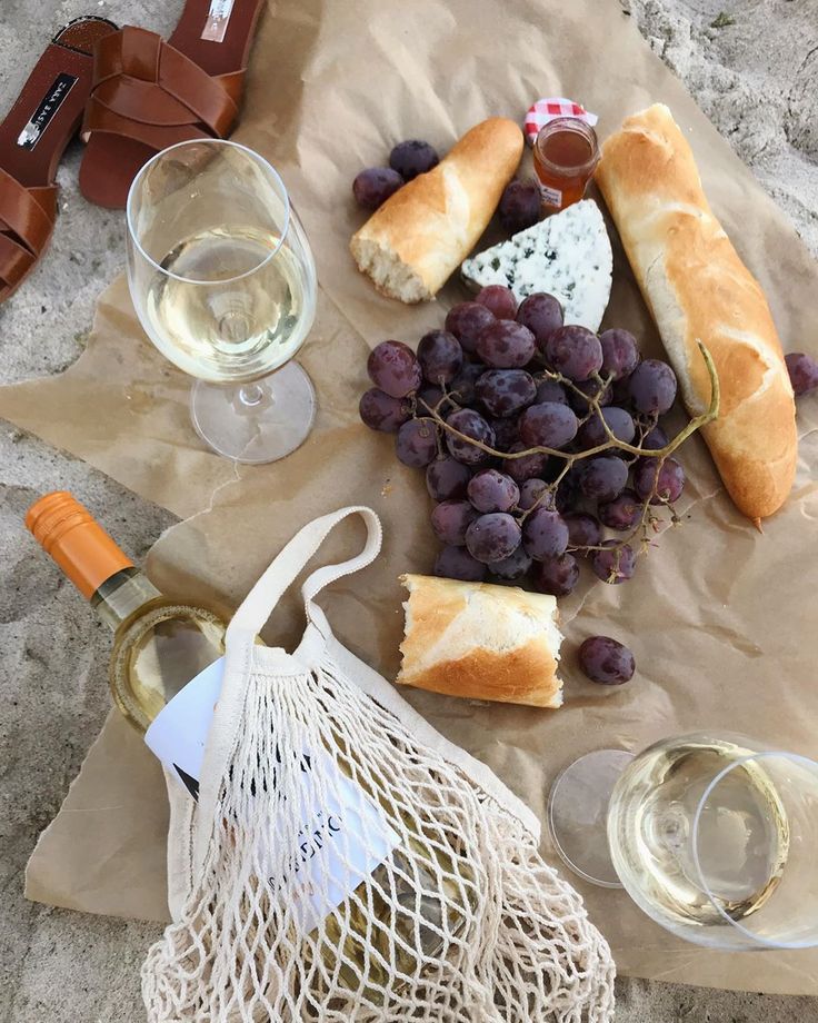 wine, bread and grapes are sitting on the sand next to a bag with a bottle of wine