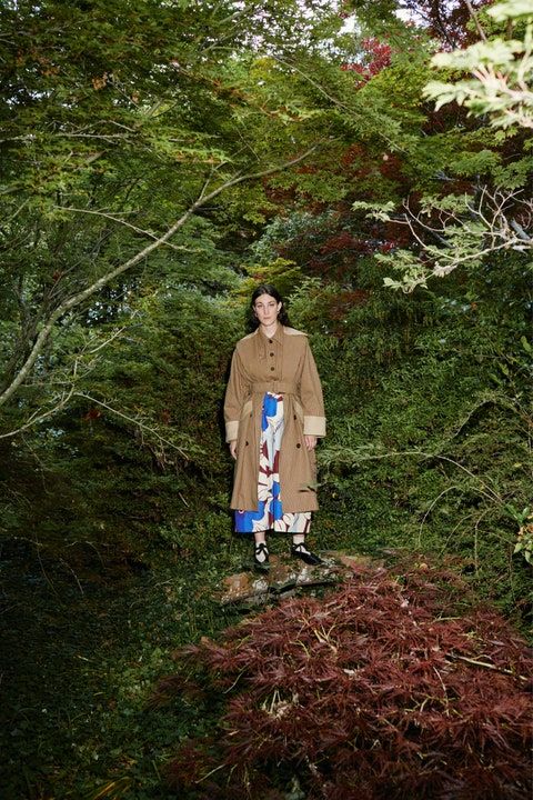 a man standing in the middle of a forest wearing a trench coat and patterned pants