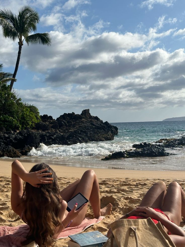 a woman laying on top of a sandy beach next to the ocean with a book in her hand