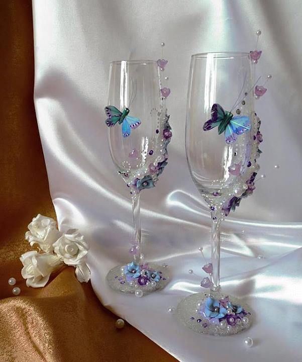 two wine glasses with butterflies on them sitting next to a white flowered bouquet and satin fabric