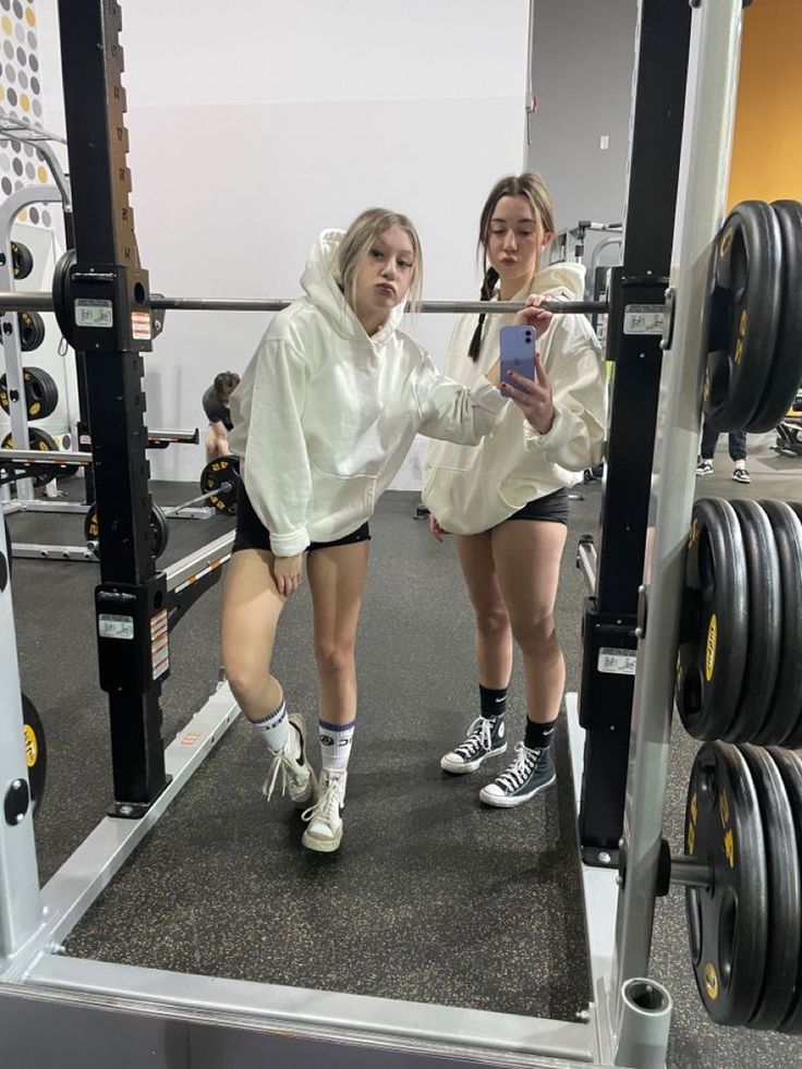 two women in white hoodies and black shorts squatting on a weight machine with one woman looking at her phone