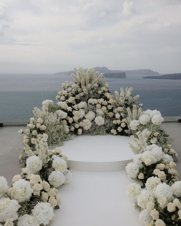 a wedding arch with white flowers and greenery on the ground next to an ocean