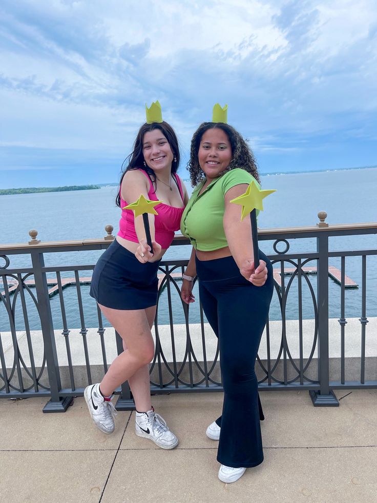 two women posing for the camera in front of a railing with water and sky behind them