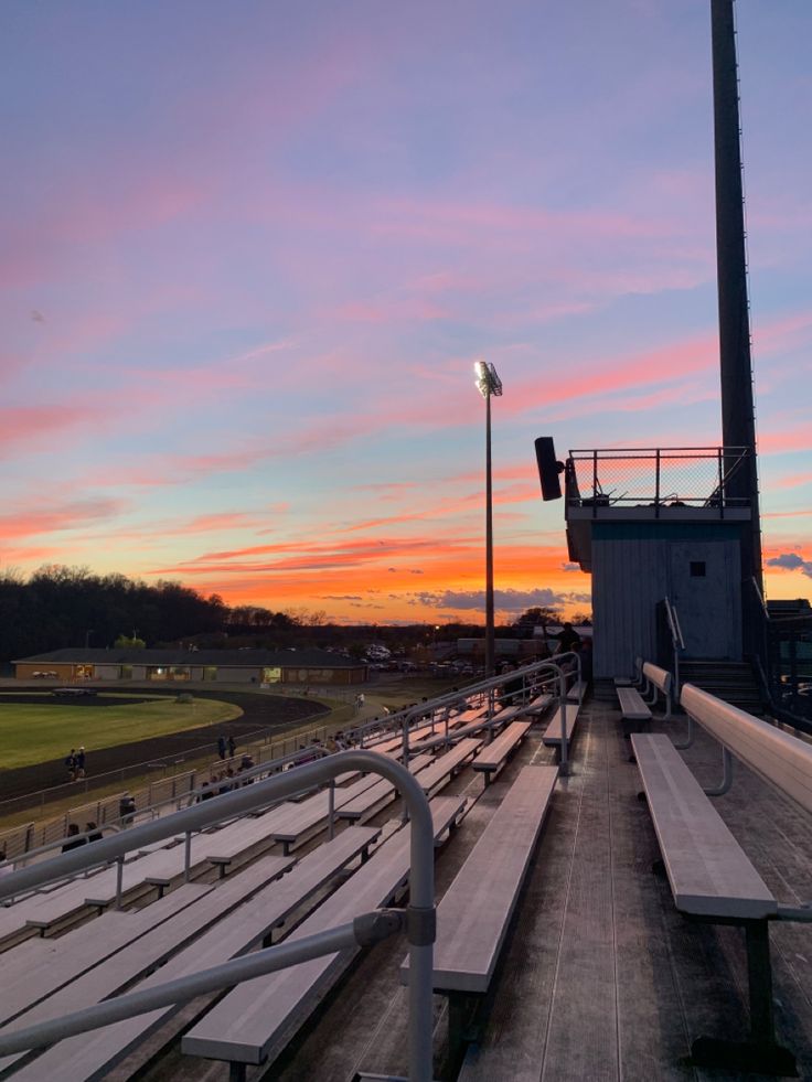 the sun is setting over an empty baseball field with benches and lights in front of it