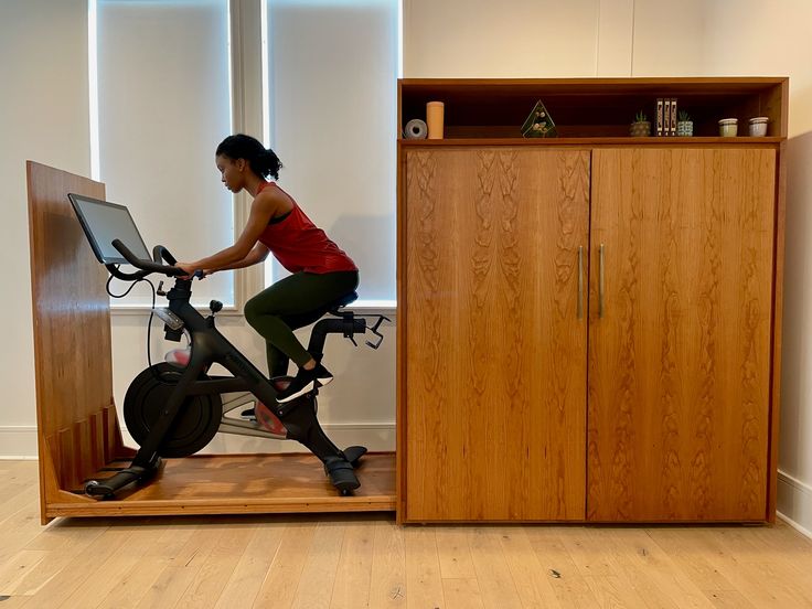 a woman on an exercise bike in front of a wooden cabinet with shelves and doors