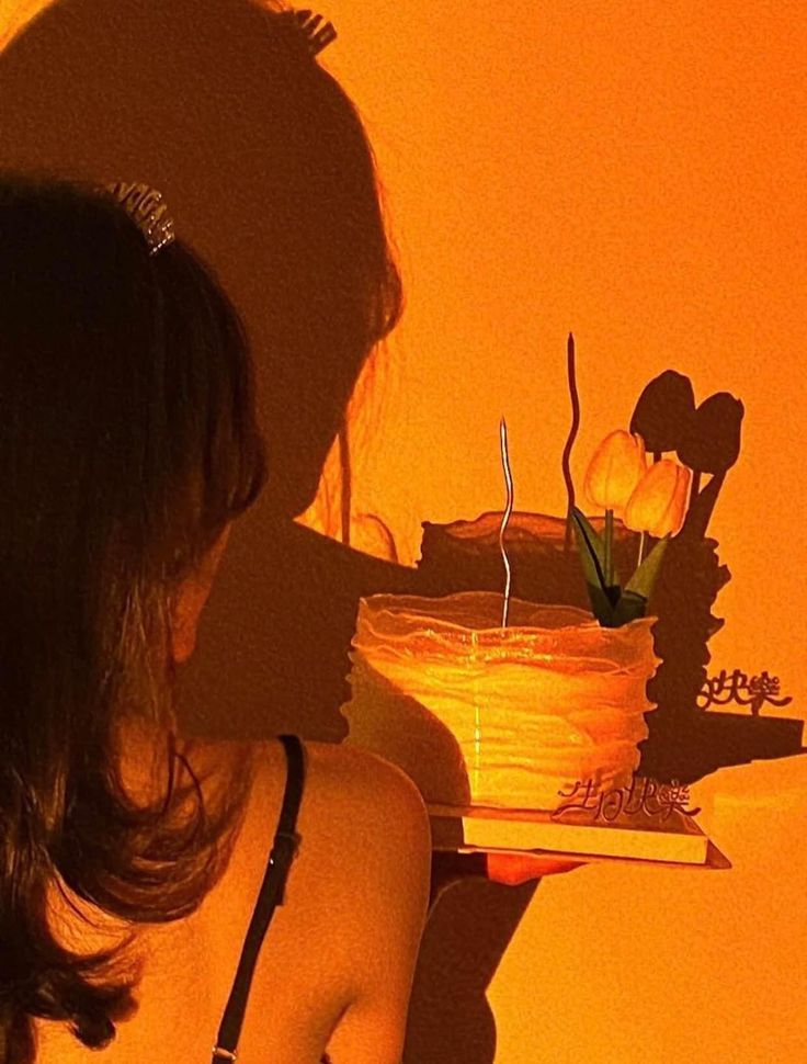 a woman holding a cake with candles on it in front of an orange wall behind her