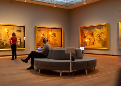 a man sitting on a couch in an art gallery looking at his cell phone and another person standing next to him