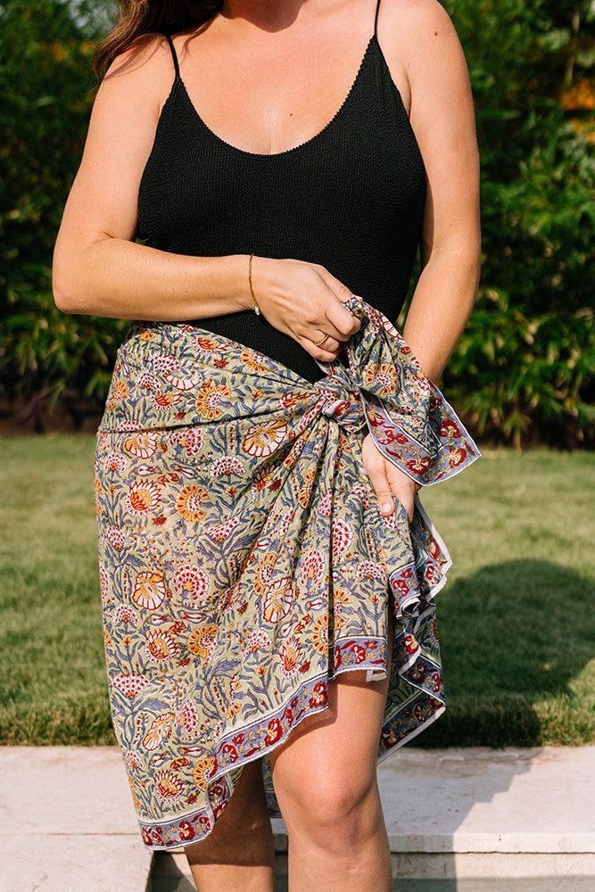 A model wears the Jaipur Block Print Sarong knotted around her waist. It is made of flowy cotton fabric with a traditional Indian block-print pattern. The fabric features a light green background with vibrant orange and red florals printed on it. The sarong has a blue floral border. Bohemian Sarong With Tropical Print For Beach Season, Summer Floral Print Sarong For Poolside, Bohemian Tropical Print Sarong For Beach Party, Floral Print Sarong For Beach Cover-up, Bohemian Patterned Sarong For Summer, Beachy Floral Print Sarong For Poolside, Beachy Sarong With Floral Print For Poolside, Beachy Poolside Sarong With Floral Print, Bohemian Tropical Print Sarong For Beach Cover-up