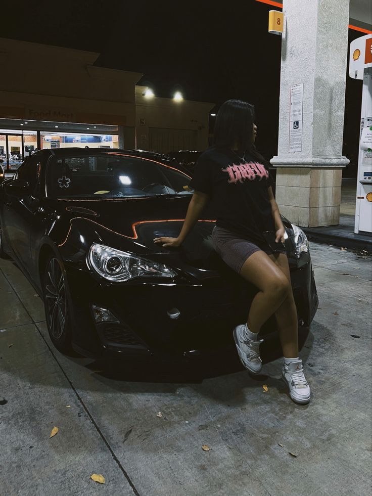 a woman sitting on the hood of a black car in front of a gas station